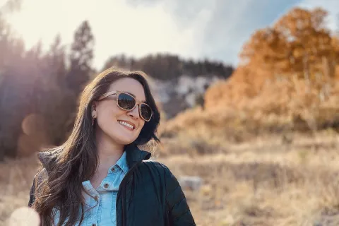 Smiling woman wearing sunglasses and jacket enjoying sunny autumn nature outdoors with trees in background