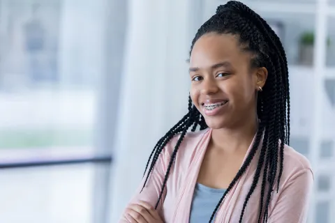 Smiling girl with braided hair and braces, standing confidently indoors with natural light.
