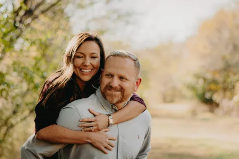 Smiling woman hugs man from behind outdoors in a bright, natural setting with fall foliage in background