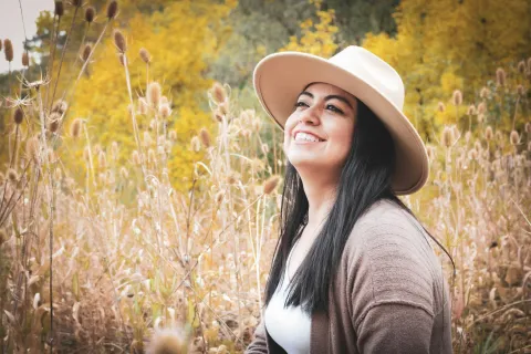 Smiling woman wearing a hat and cardigan sitting in a field with autumn foliage in the background