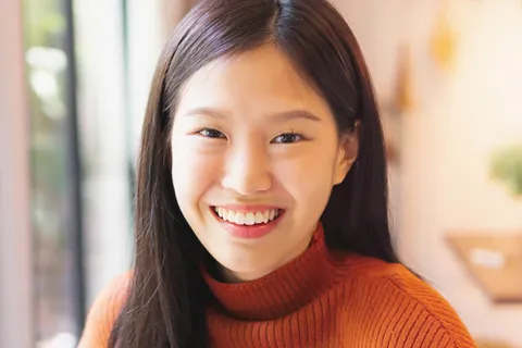 Smiling young woman wearing an orange sweater in a cozy, well-lit indoor setting.