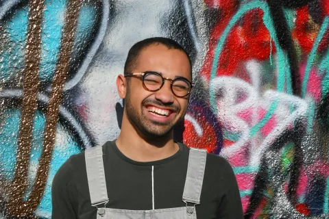 Smiling man in glasses wearing grey overalls and holding a black cap standing against colorful graffiti wall.