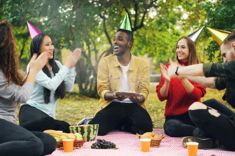 Group of friends wearing party hats celebrating a birthday at an outdoor picnic with cake and snacks