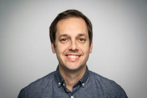 Smiling man with short brown hair wearing a blue button-up shirt against a light gray background