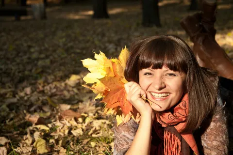 Smiling woman lying on autumn leaves holding bright yellow and orange maple leaves in a sunny park.