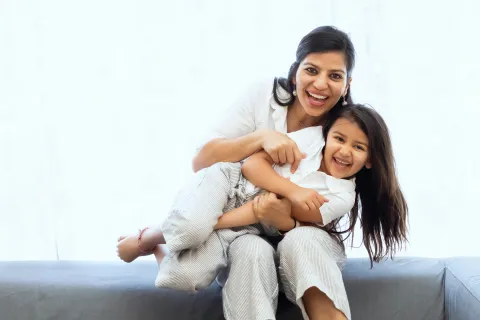 Mother and daughter smiling and hugging each other while sitting on a gray couch in bright room.