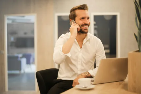 Smiling man in white shirt using phone and laptop at wooden desk with coffee cup in modern office space