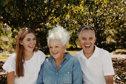 Three generations smiling and laughing outdoors under leafy trees on a sunny day.