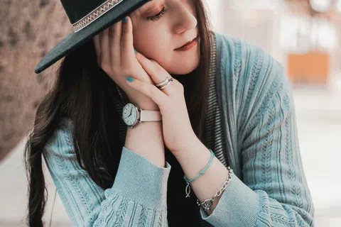 Young woman in a black hat and gray sweater sitting with eyes closed and hands resting on her face.