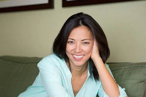 Smiling woman with dark hair in light blue blouse relaxing on green couch in cozy living room.