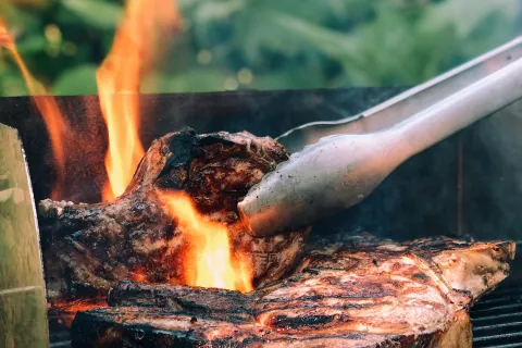 Close-up of thick steaks grilling over hot flames on a charcoal barbecue with tongs flipping the meat.