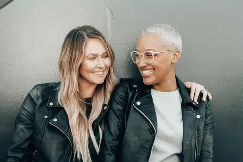 Two women in leather jackets smiling and embracing each other against a gray wall background.