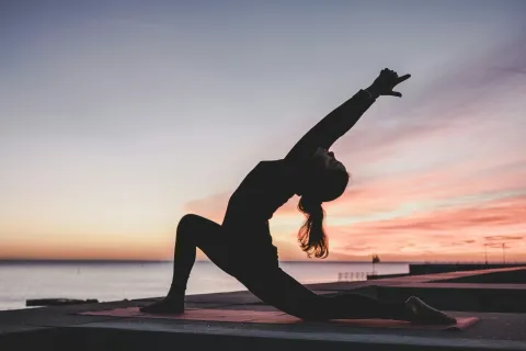 Silhouette of a woman practicing yoga in a deep stretch pose at sunset by the ocean on a mat.