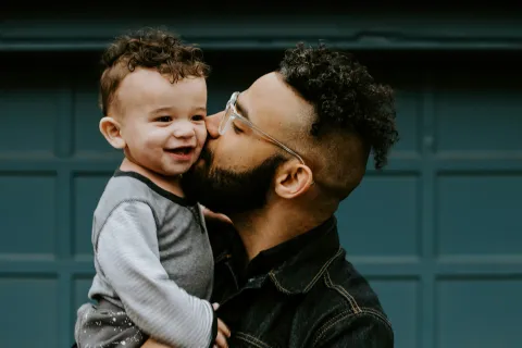Father wearing glasses kisses smiling toddler with curly hair in front of blue garage door.