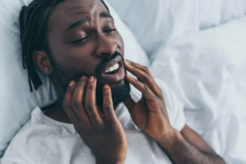 A man experiencing discomfort while lying in bed, holding his face with both hands.