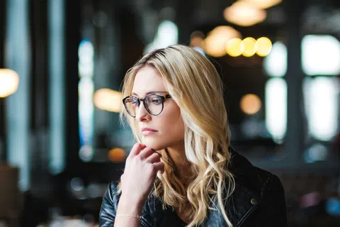 Blonde woman in glasses and black leather jacket thoughtfully posing in dimly lit modern cafe interior.