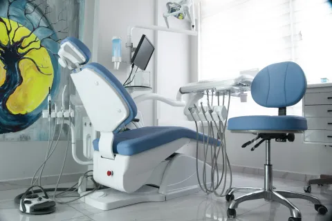 Modern dental clinic equipped with blue dental chair, tools, monitor, and artistic wall decor in bright room.