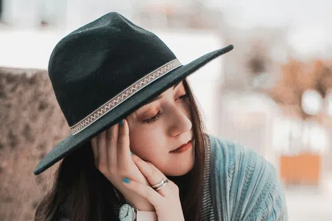 Young woman in a black hat and light blue sweater sitting with eyes closed, resting her face on her hands.