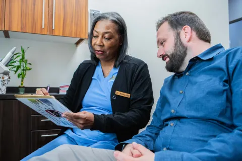 Female doctor in blue scrubs explaining medical information to male patient in clinic office.