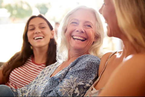 Three women of different ages happily smiling and conversing in a bright, casual setting.