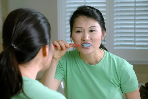 Woman brushing teeth with a red toothbrush in front of a mirror wearing a green shirt indoors.
