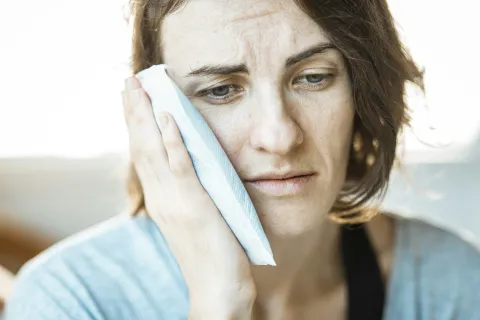 Woman holding an ice pack to her cheek looking distressed with pain and discomfort