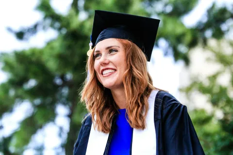 Smiling female graduate in cap and gown outdoors with blurred green trees in background