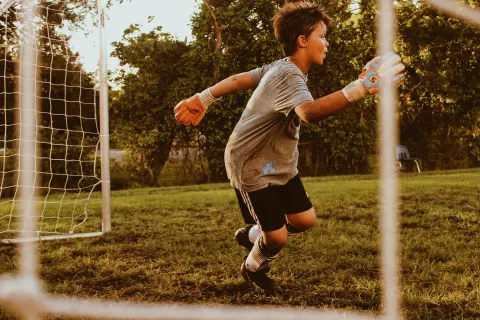 Young boy goalkeeper in gray shirt and gloves ready to block a soccer ball in goal during outdoor game.