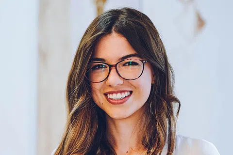 A smiling woman with long brown hair, wearing glasses and a white blouse in a bright indoor setting.