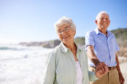 Happy elderly couple holding hands at the beach with a sunny sky and gentle waves in the background.