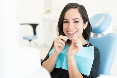 Smiling woman in a dental chair holding clear aligners during a dental appointment.