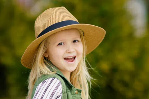 Smiling young girl wearing a tan hat and green vest standing outdoors with blurred greenery background.