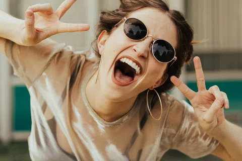 Young woman in sunglasses and hoop earrings smiling widely and making peace signs outdoors