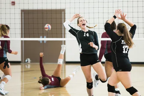 Female volleyball players celebrating a point during an indoor match with a player on the floor in the background.