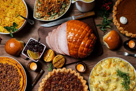 Thanksgiving feast with glazed ham, pecan pie, pumpkin pie, casseroles, dinner rolls, gravy, and candles on wooden table.