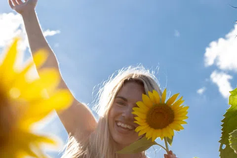 Smiling woman in striped dress holding a sunflower in a sunny sunflower field under a blue sky with clouds