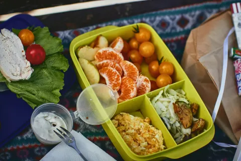 Yellow lunchbox with tangerine slices, cherry tomatoes, scrambled eggs, and vegetables on a picnic blanket.