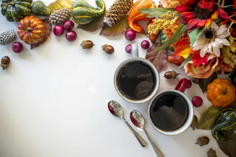 Two cups of coffee surrounded by autumn leaves, pumpkins, pinecones, acorns, and decorative flowers on white.