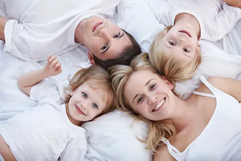 Happy family of four lying on bed wearing white clothes, smiling and looking up at the camera.