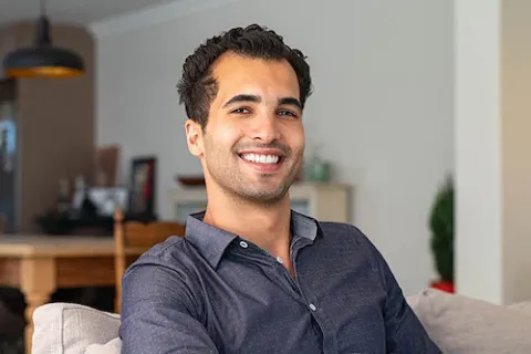 Smiling young man with dark hair and gray shirt sitting on a couch in a modern living room.