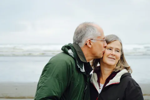 Older man in green jacket kisses smiling woman in black jacket at the beach on a cloudy day.