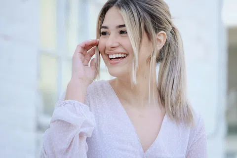 Smiling young woman with blonde hair in a ponytail wearing a light pink blouse outdoors