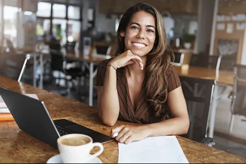 Smiling woman sitting at a wooden table with a laptop coffee cup and documents in a bright office space