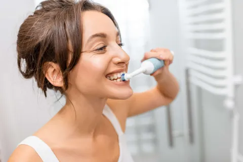 Woman brushing her teeth with an electric toothbrush in a bright bathroom setting.