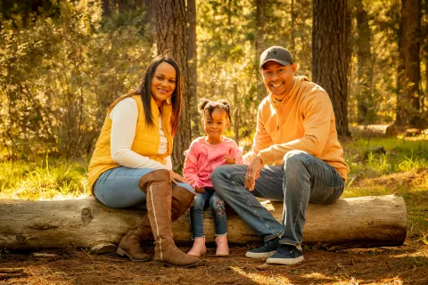 Smiling family of three sitting on a log in a sunlit forest during autumn outdoor outing