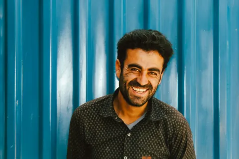Smiling man with dark hair and beard standing in front of blue corrugated metal wall wearing a patterned shirt.