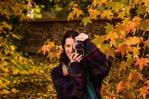 Woman in autumn sweater holding a camera amidst vibrant fall leaves in a forest setting.