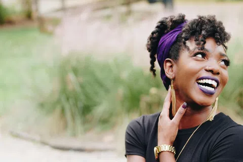 Smiling young woman with purple headband, gold earrings, and lipstick wearing black shirt outdoors