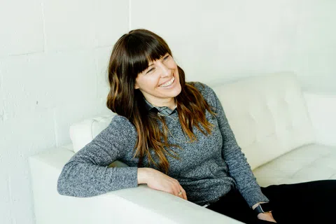 Smiling woman with brown hair wearing gray sweater sitting relaxed on white couch against white wall.