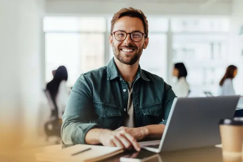 Smiling man with glasses working on a laptop at a modern office desk with blurred coworkers in background
